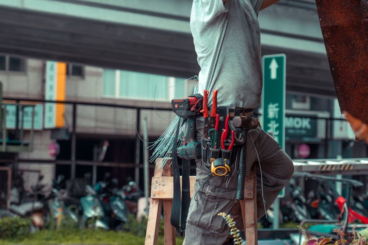 services-03 Handyman standing on a ladder with tool belt, repairing outdoors with visible tools.