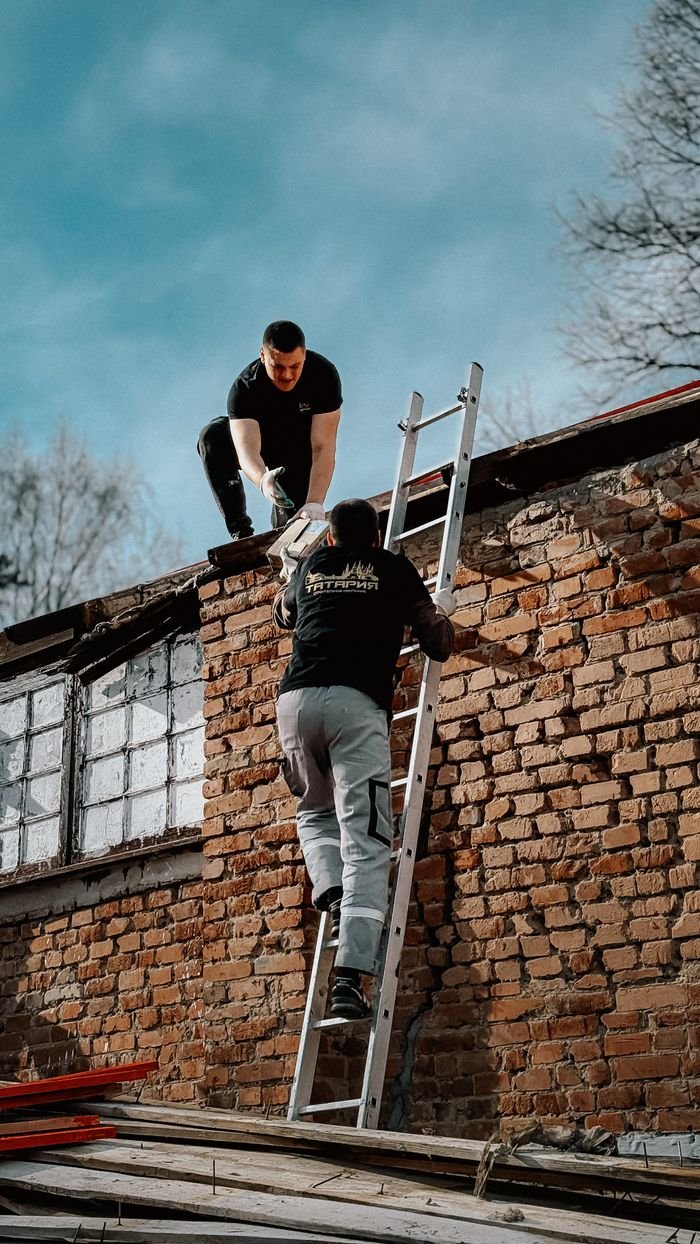 our-vission Two construction workers using a ladder to reach a brick building roof under a blue sky.