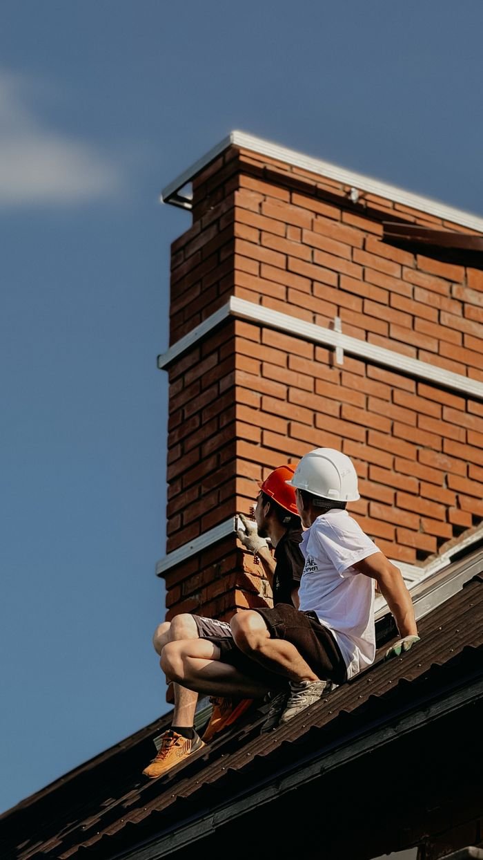 our-vission Two construction workers with hard hats repairing a chimney on a house roof under a clear sky.