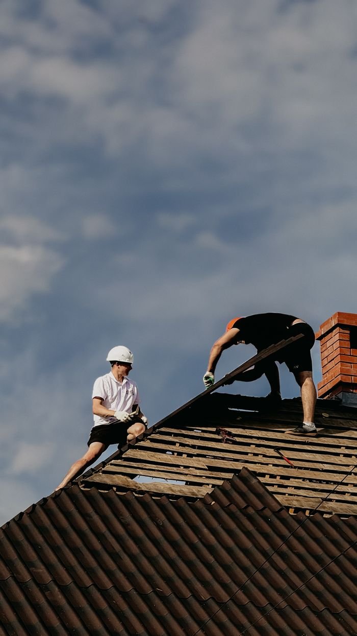 behind-the-scene-04 Two roofers working on a roof repair with a clear blue sky, demonstrating teamwork and construction skills.